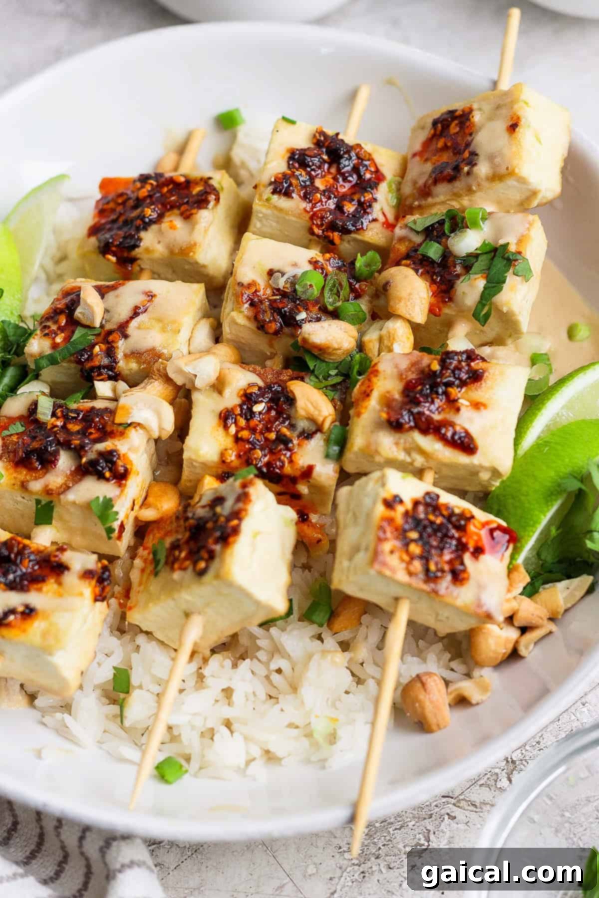 Skewered tofu cubes with chili sauce are placed over a bed of rice, garnished with chopped green onions, peanuts, and lime wedges.