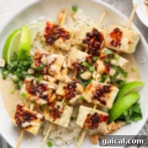 A dish with skewered tofu topped with chili sauce on rice, garnished with lime wedges, green onions, peanuts, and herbs.