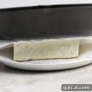 A block of tofu being pressed between a skillet and white plate, with paper towels underneath.