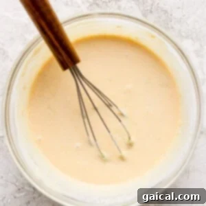 A glass bowl filled with a light beige batter. A whisk with a wooden handle rests in the batter. The surface beneath the bowl is textured and light-colored.