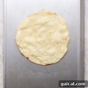 A round, uncooked pizza dough is placed on a metal baking sheet with a textured surface.
