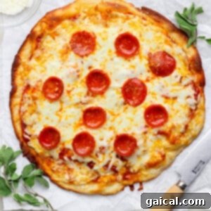 A pepperoni pizza on parchment paper with a knife beside it, garnished with herbs. A small bowl of grated cheese is in the top-left corner.