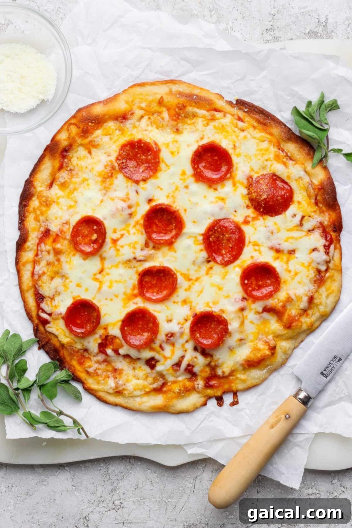 Rapid Four Ingredient Protein Pizza Crust 5 A pepperoni pizza on parchment paper with a knife beside it, garnished with herbs. A small bowl of grated cheese is in the top-left corner.