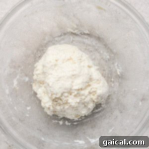 A ball of dough in a clear glass bowl on a light textured surface.