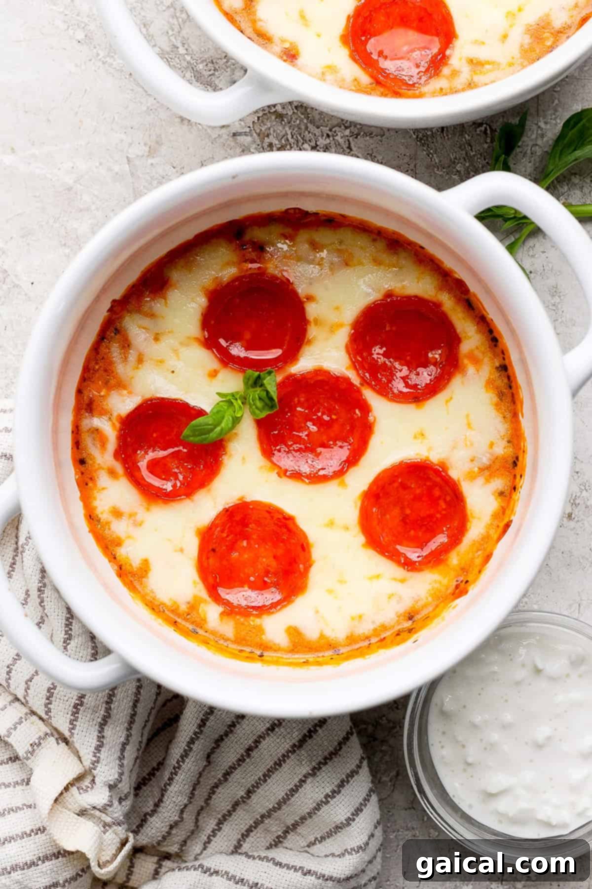 Personal Cottage Cheese Pizza Bowl 2 A white bowl filled with baked pepperoni and cheese dip, garnished with a basil leaf, placed on a cloth next to a small bowl of dipping sauce.