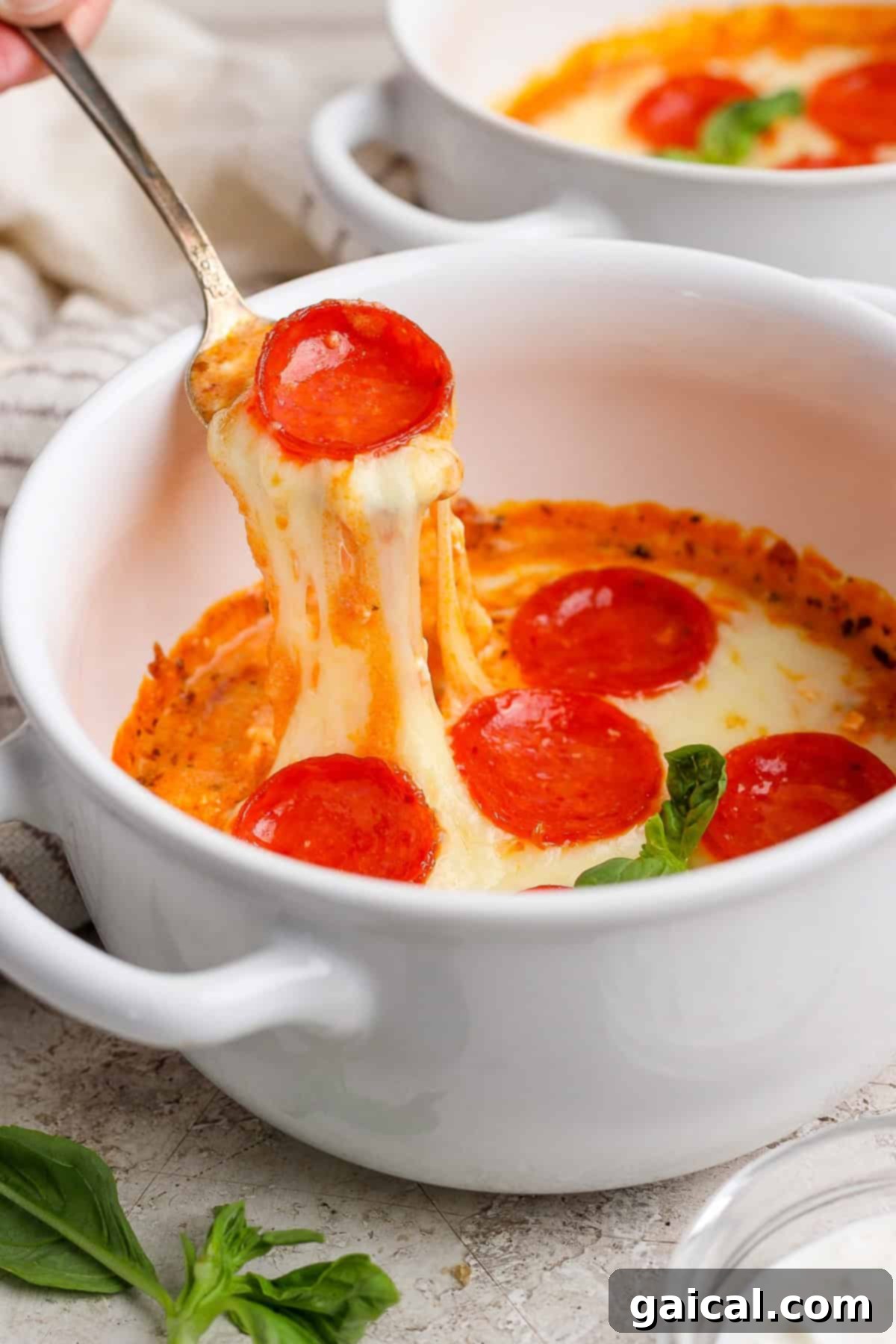 Personal Cottage Cheese Pizza Bowl 8 A spoon lifts cheesy pepperoni pizza dip from a white bowl. Fresh basil leaves are visible nearby.