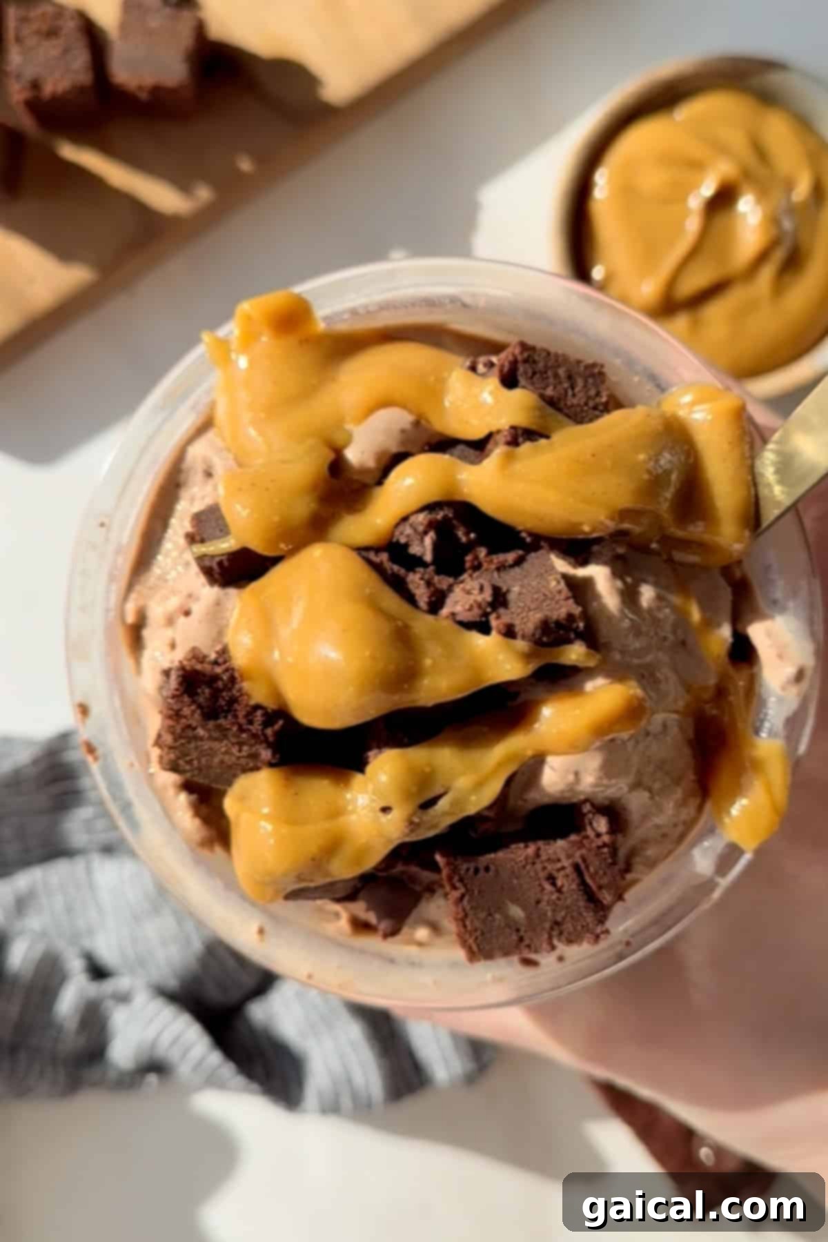 Decadent Chocolate Brownie Protein Creami Ice Cream 2 A cup of rich chocolate ice cream, generously topped with a golden caramel sauce and several chunks of delicious brownie. A spoon is gently resting in the ice cream, inviting a taste. In the blurred background, a bowl of more caramel sauce hints at the sweetness to come.