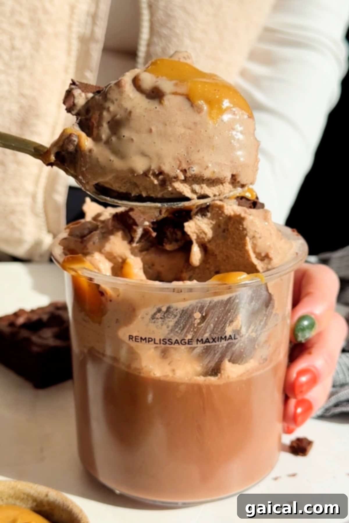 Decadent Chocolate Brownie Protein Creami Ice Cream 4 A person holds a spoon brimming with delicious ice cream, drizzled with caramel sauce, over a clear cup. The cup, filled to its maximum line, showcases the rich texture of the ice cream and its brownie chunks. A homemade brownie is visible in the soft background.