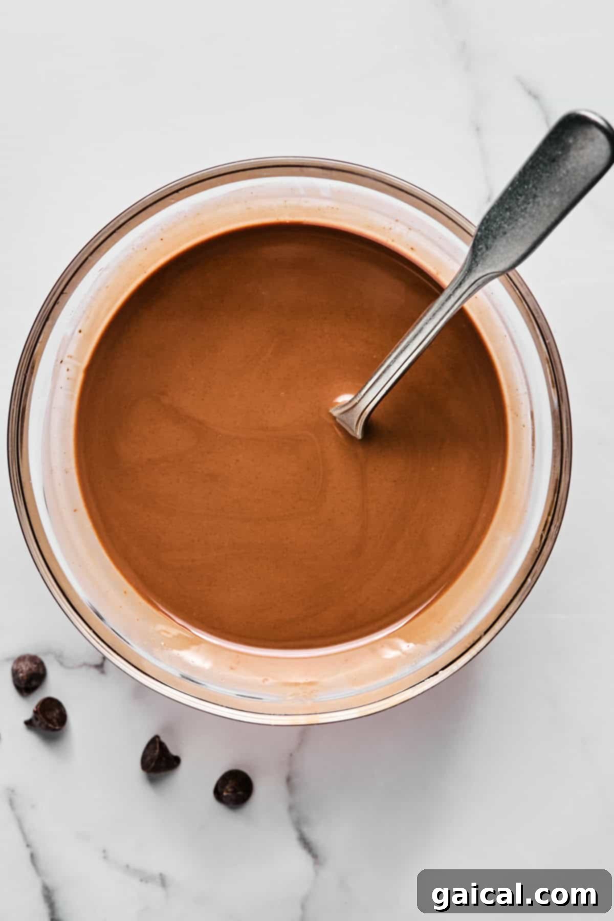 Create Your Own Magical Chocolate Coating 4 Bowl of melted chocolate with a metal spoon, placed on a white marble surface next to a few chocolate chips.