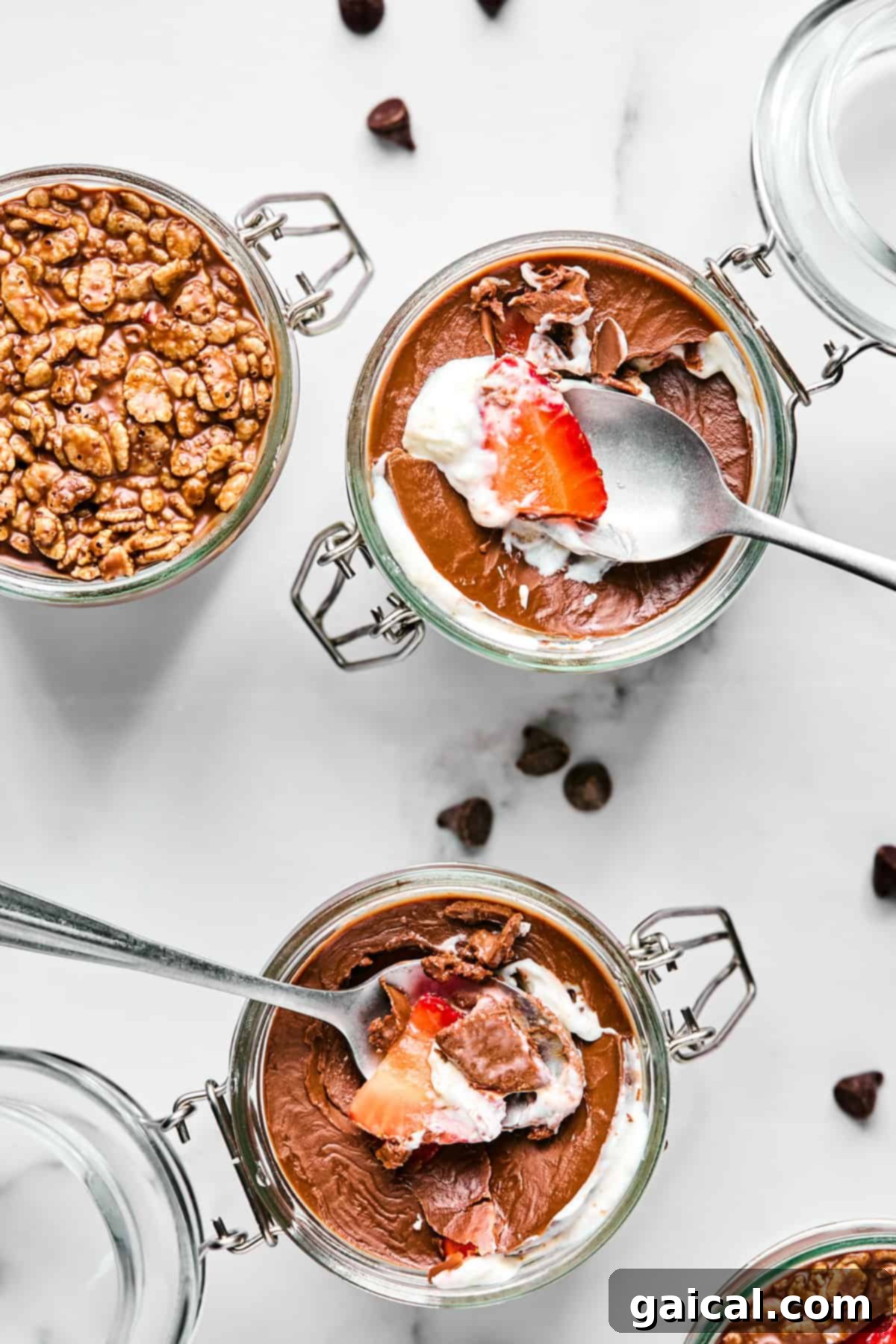 Create Your Own Magical Chocolate Coating 5 Three glass jars of chocolate mousse topped with whipped cream, strawberries, and cereal, with spoons inside. A few chocolate chips are scattered on the white surface.