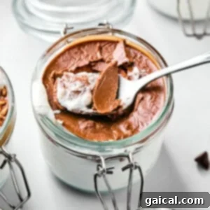 A spoon scoops chocolate dessert from a glass jar, revealing a creamy white layer underneath. Another jar with a similar dessert is partially visible in the background.