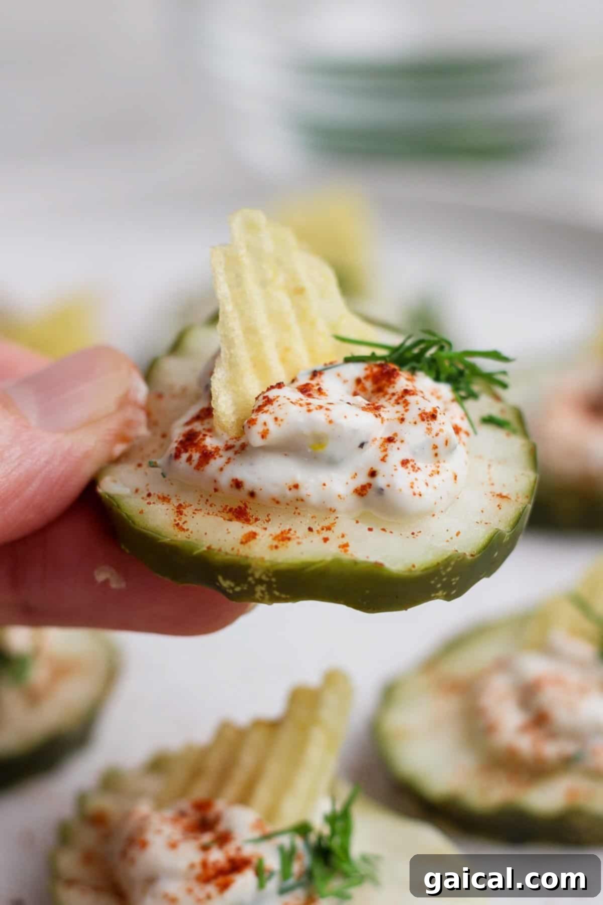 A hand holding a deviled pickle, topped with creamy filling, paprika, a piece of ridged potato chip, and a sprig of dill.