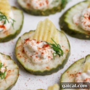 A close-up of a plate of Easy Deviled Pickles, showing cucumber slices topped with creamy filling, a potato chip, fresh dill, and paprika on a white surface.