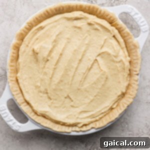 An unbaked pie crust, filled with a rich, creamy batter, sits patiently in a pristine white baking dish on a subtly textured surface, awaiting its culinary transformation.