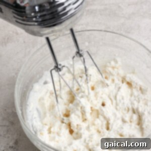 An electric mixer actively whips cream to a fluffy consistency in a clear glass bowl, placed on a gleaming light-colored kitchen counter, showcasing the process of preparing a rich topping.