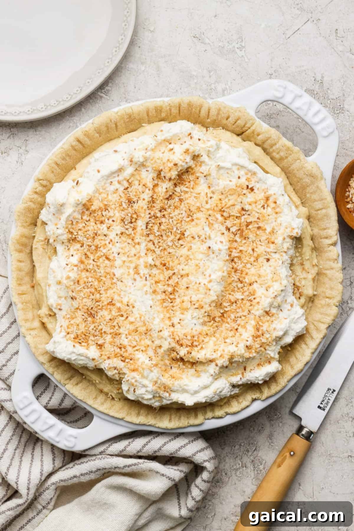 Luscious Coconut Cream Pie 5 A beautifully crafted coconut cream pie, generously adorned with swirls of whipped cream and a sprinkle of golden toasted coconut flakes, resting elegantly on a light-colored surface. Nearby, a sharp knife and a stylish striped cloth hint at the joy of serving this tropical delight.