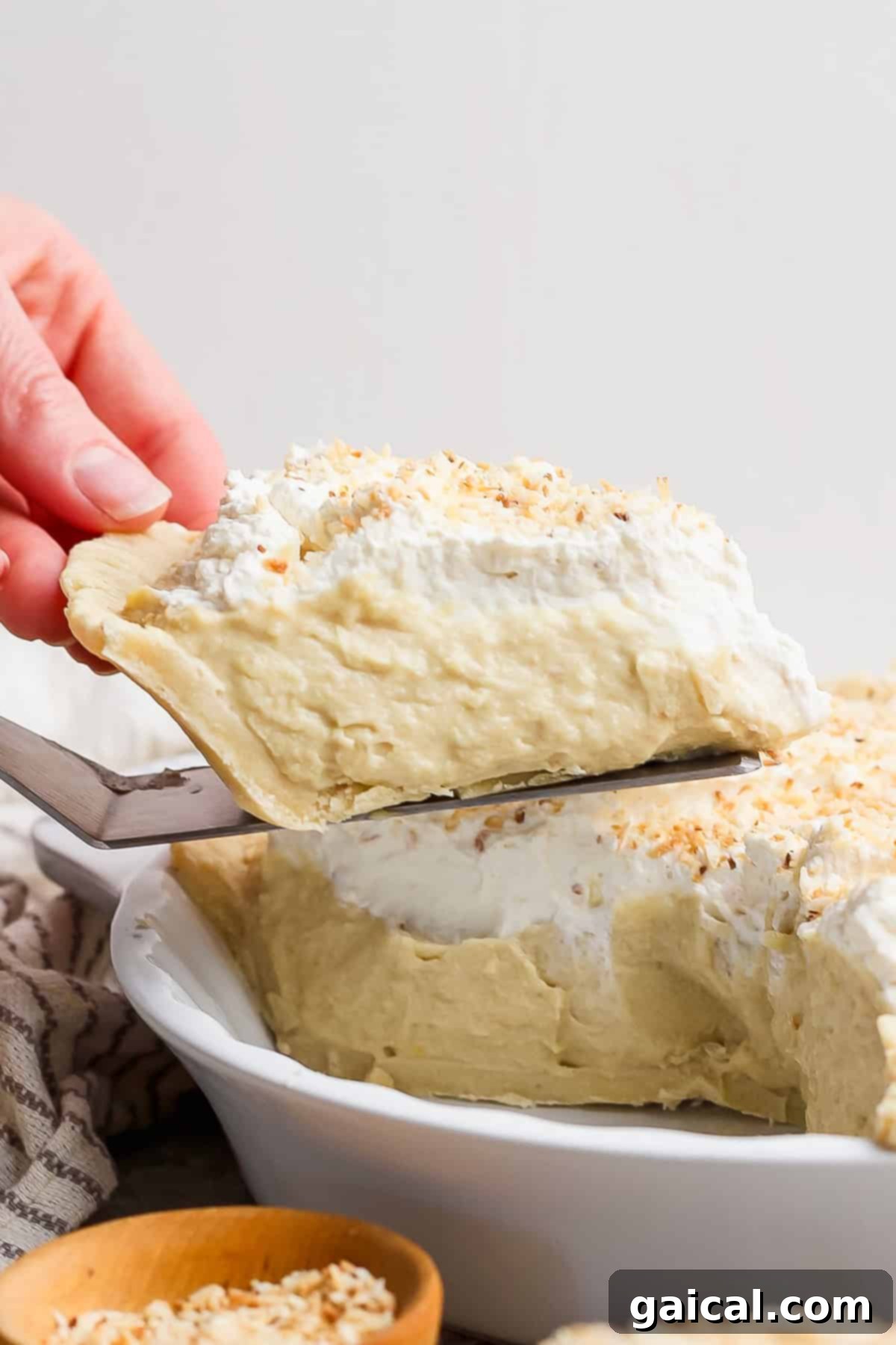 Luscious Coconut Cream Pie 6 A hand gently serves a generous slice of creamy coconut pie, topped with a dollop of fluffy whipped cream and a delicate sprinkle of toasted coconut, from a pristine white pie dish, capturing a moment of dessert indulgence.