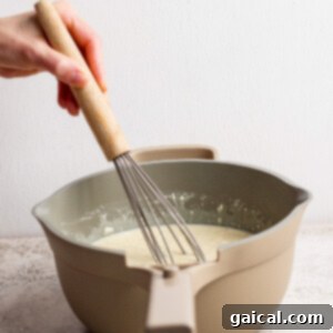 A person expertly whisks a creamy batter in a beige mixing bowl, set on a bright, light-colored countertop, creating a smooth and consistent mixture for the pie filling.