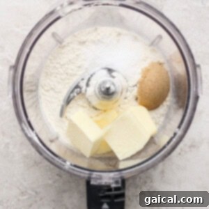 The food processor bowl contains a mixture of flour, brown sugar, salt, and butter, representing the initial steps of pie crust preparation, resting on a clean, light surface.