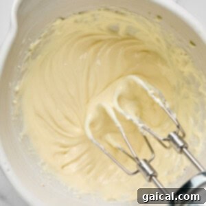 Protein-Packed Carrot Cake Roll 14 Electric mixer beaters in a bowl with creamy batter on a marble countertop.