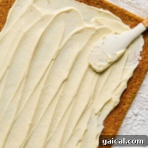 Protein-Packed Carrot Cake Roll 15 A rectangular cake layer being spread with cream cheese frosting using a spatula on a floured surface.