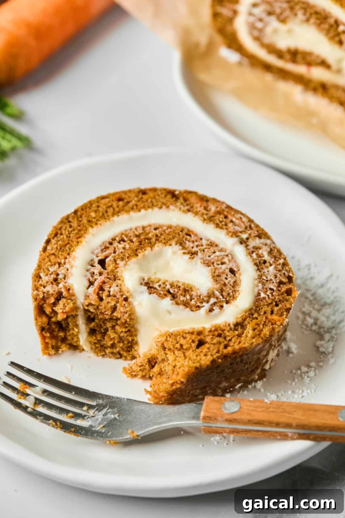 Protein-Packed Carrot Cake Roll 5 A perfect slice of Protein Carrot Cake Roll with its inviting cream cheese filling, elegantly presented on a white plate next to a fork. A larger portion of the roll and a fresh carrot are subtly blurred in the background.