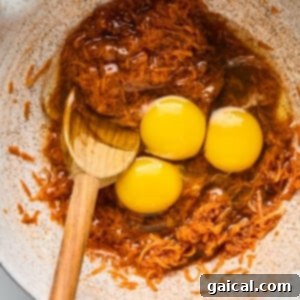 Protein-Packed Carrot Cake Roll 9 Three raw eggs are cracked into a mixing bowl containing brown shredded ingredients, with a wooden spoon partially submerged in the mixture.