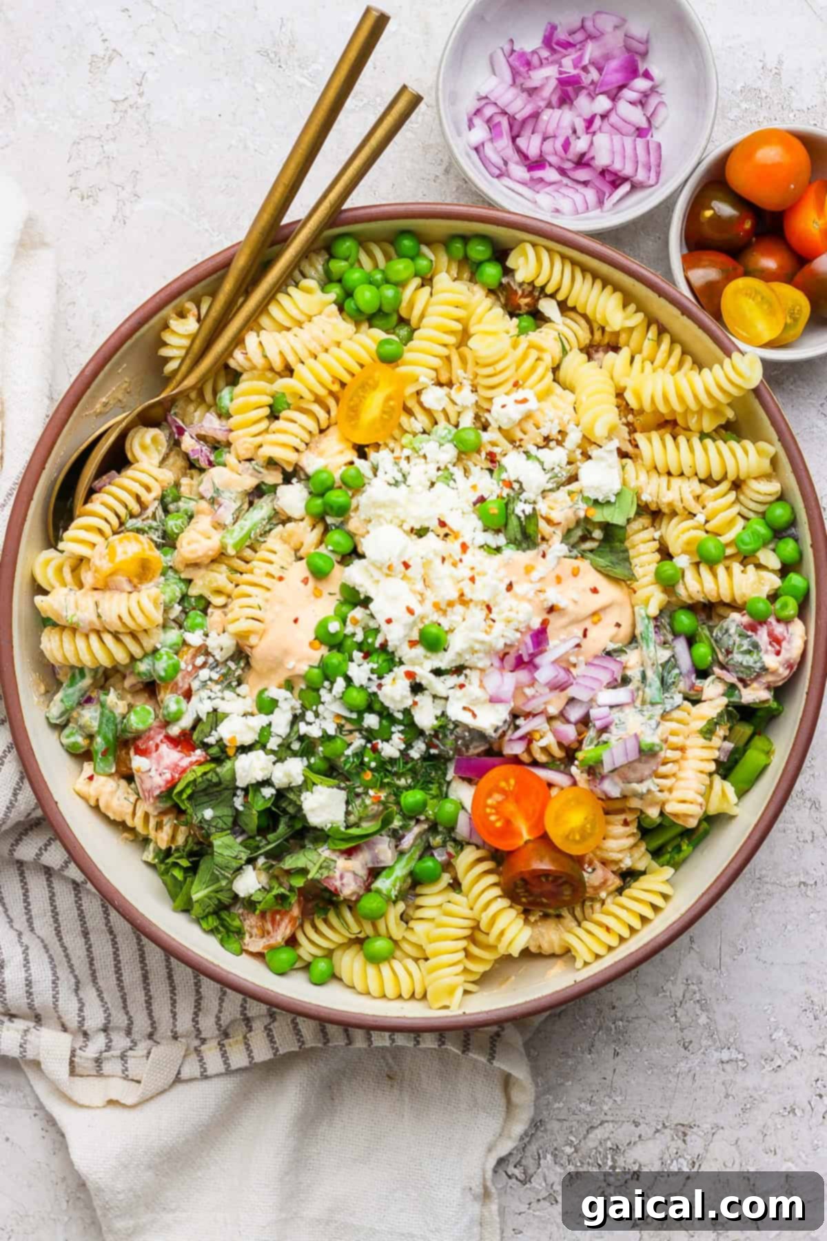 Creamy Roasted Red Pepper Pasta Delight 2 A bowl of pasta salad with fusilli, cherry tomatoes, peas, feta cheese, and red onion. Two gold utensils rest on the side. Small bowls of red onions and cherry tomatoes are nearby.