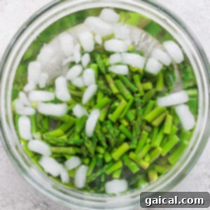 Chopped asparagus in a glass bowl of ice water, with ice cubes floating on top, against a textured white background.