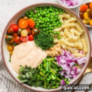 A bowl with rotini pasta, cherry tomatoes, peas, chopped red onion, green beans, fresh herbs, and a creamy sauce. A small bowl of onions and cutlery are beside it.