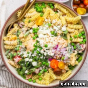 A bowl of pasta salad with fusilli, cherry tomatoes, peas, feta cheese, and red onion. Two gold utensils rest on the side. Small bowls of red onions and cherry tomatoes are nearby.