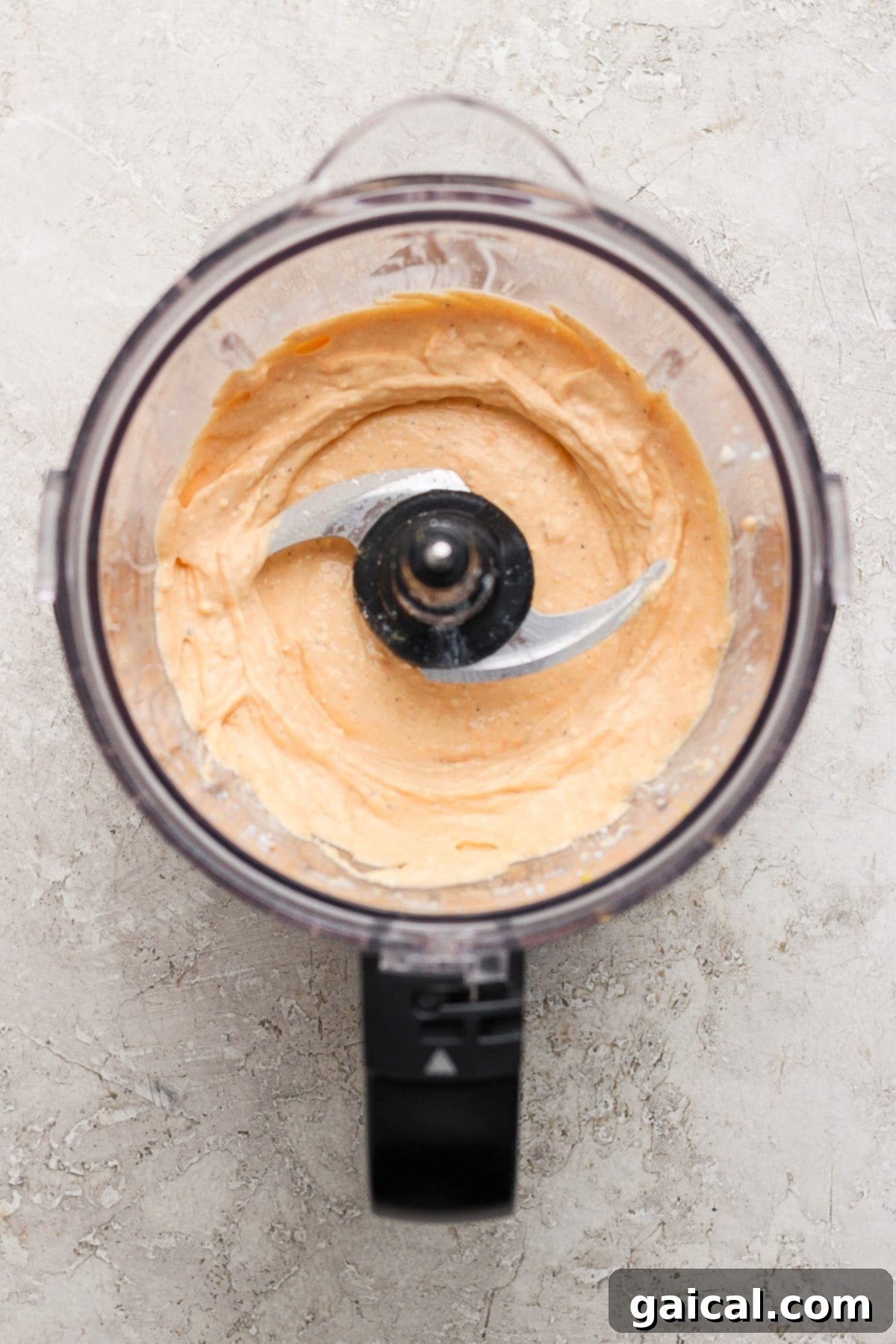 Creamy Roasted Red Pepper Pasta Delight 4 Food processor with creamy orange hummus on a gray textured surface.