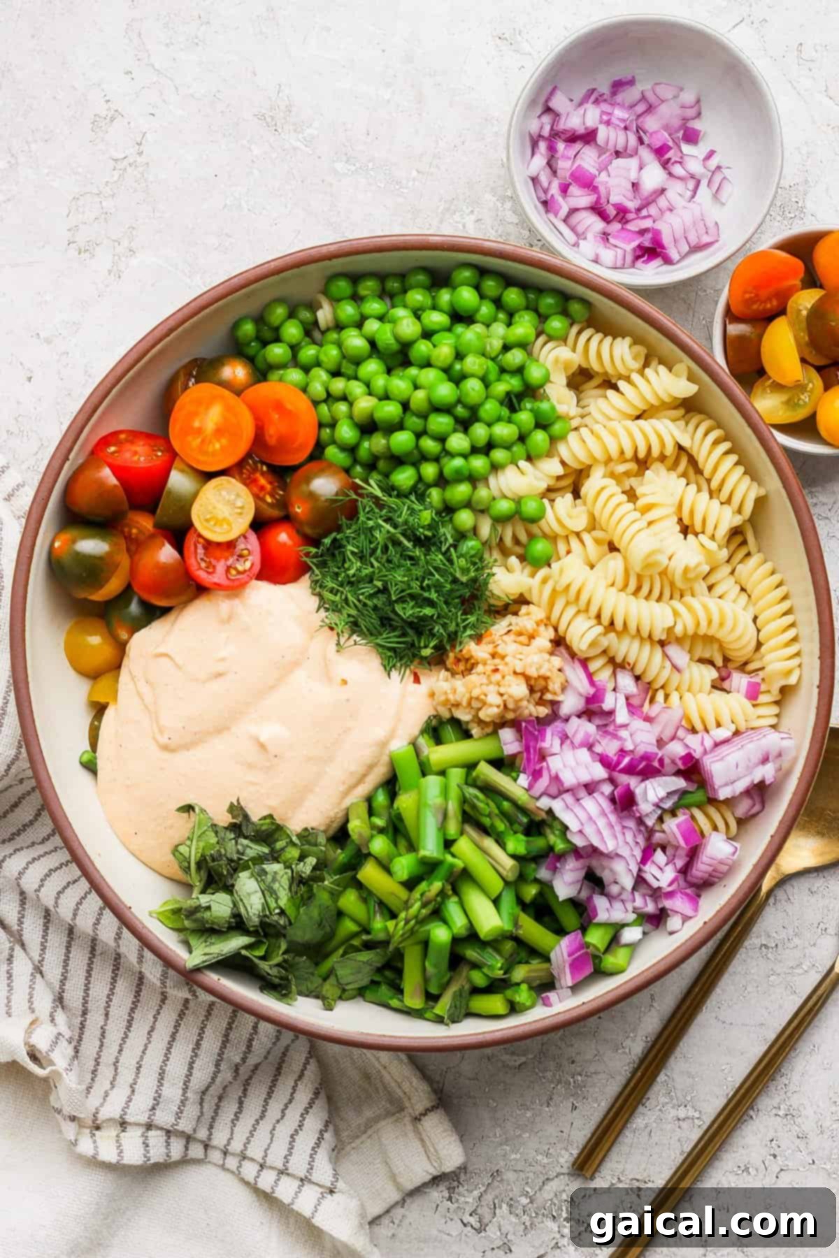 Creamy Roasted Red Pepper Pasta Delight 5 A bowl with rotini pasta, cherry tomatoes, peas, chopped red onion, green beans, fresh herbs, and a creamy sauce. A small bowl of onions and cutlery are beside it.