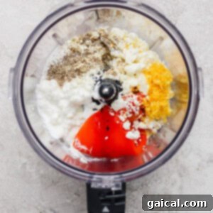 Ingredients including pepper, feta cheese, cottage cheese, lemon zest, and red peppers in a food processor, ready to be blended.
