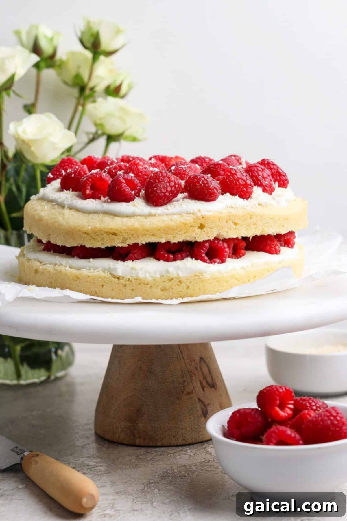 Layered Coconut Bliss with Fresh Raspberries 2 Two-layer coconut cake with white frosting and vibrant red raspberries on top, elegantly displayed on a wooden cake stand. White flowers and a bowl of fresh raspberries adorn the background, emphasizing the cake's beauty.