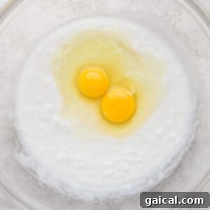 Layered Coconut Bliss with Fresh Raspberries 6 Two raw eggs cracked on top of a creamy mixture in a clear glass bowl on a textured surface, ready to be incorporated into the cake batter.