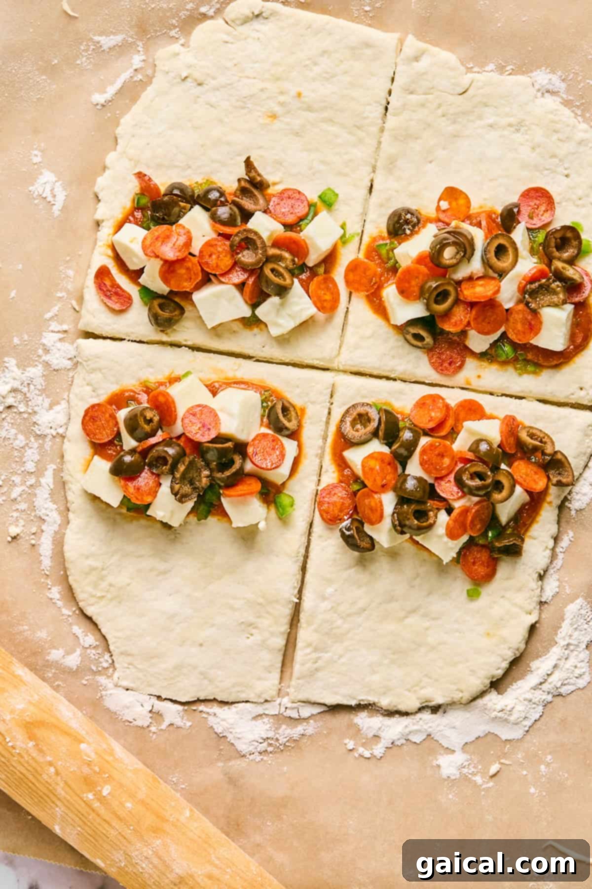 Four sections of raw dough, resembling pizza hot pockets, are topped with cheese cubes, pepperoni slices, olives, and green onions on a floured surface.