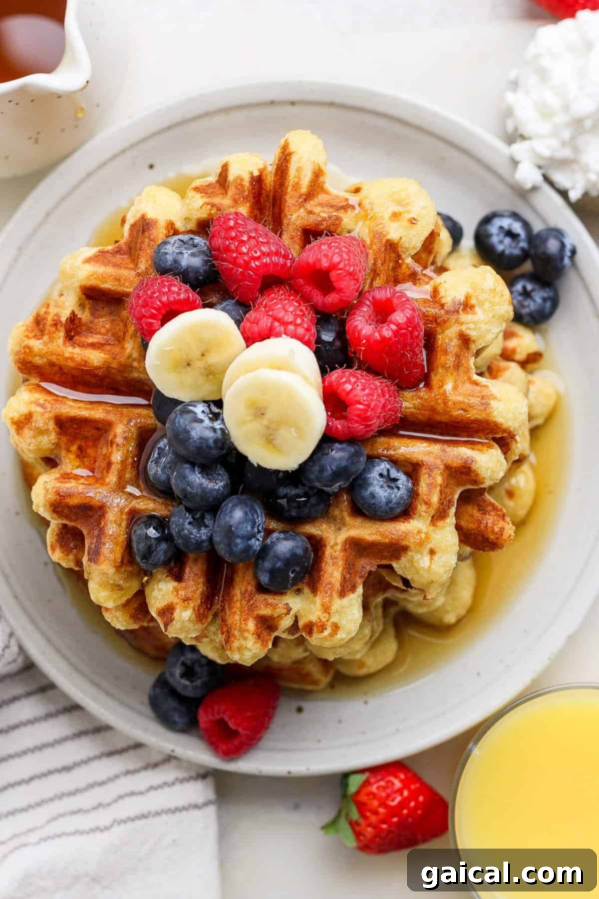 A stack of golden-brown cottage cheese waffles, generously topped with vibrant blueberries, sweet raspberries, and thinly sliced bananas, elegantly arranged on a white plate. A refreshing glass of orange juice and a neatly striped napkin complete the inviting breakfast scene.