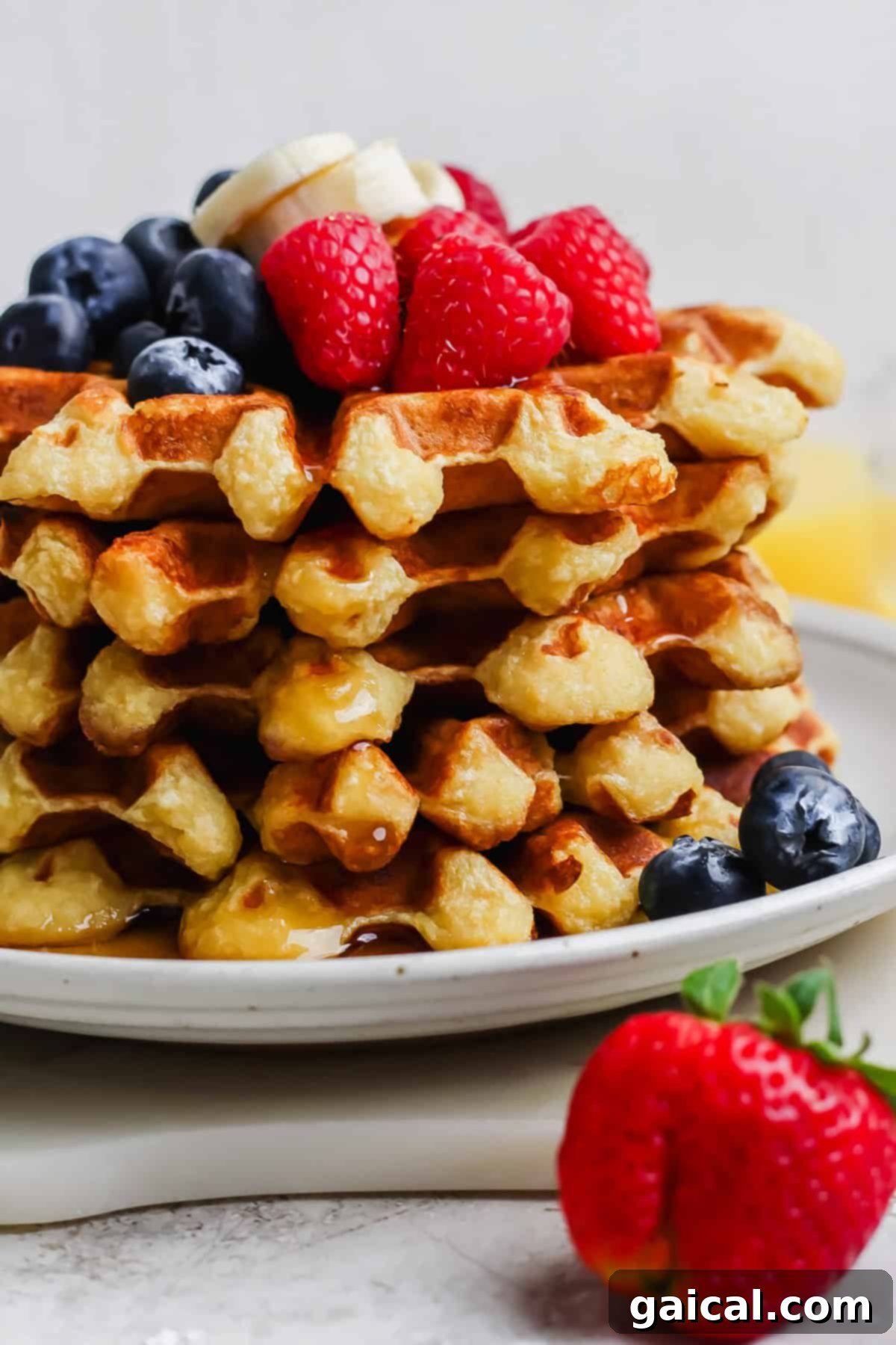 A beautifully arranged stack of golden-brown waffles, generously adorned with fresh banana slices, vibrant raspberries, and plump blueberries, all glistening with a drizzle of sweet syrup. A single, juicy strawberry sits invitingly on the edge of the white plate, ready to be enjoyed.