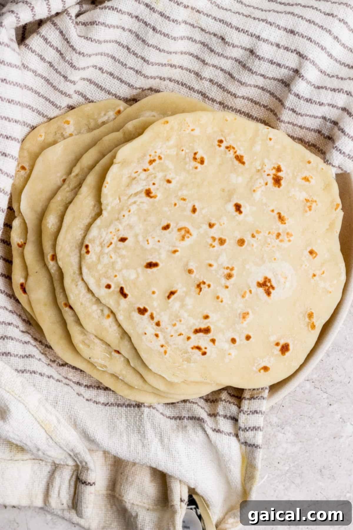 A beautifully arranged stack of golden-brown homemade flatbreads, indicating their soft and pliable texture, resting on a clean, striped kitchen towel.