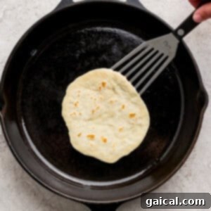 A hand uses a spatula to lift a tortilla actively cooking and bubbling in a black cast iron skillet, on a light-colored kitchen surface, showing the browning process.
