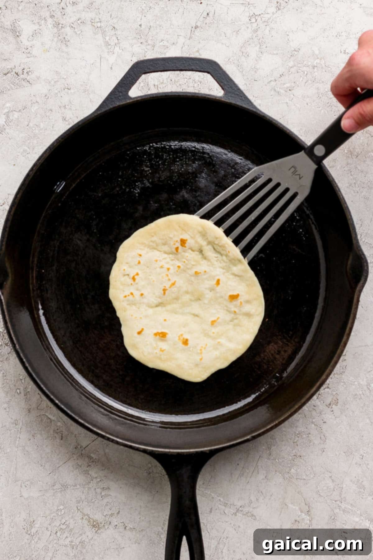 A hand expertly uses a spatula to lift a freshly cooking tortilla from a black cast iron skillet, revealing its lightly browned and bubbly surface on a light-colored kitchen counter.
