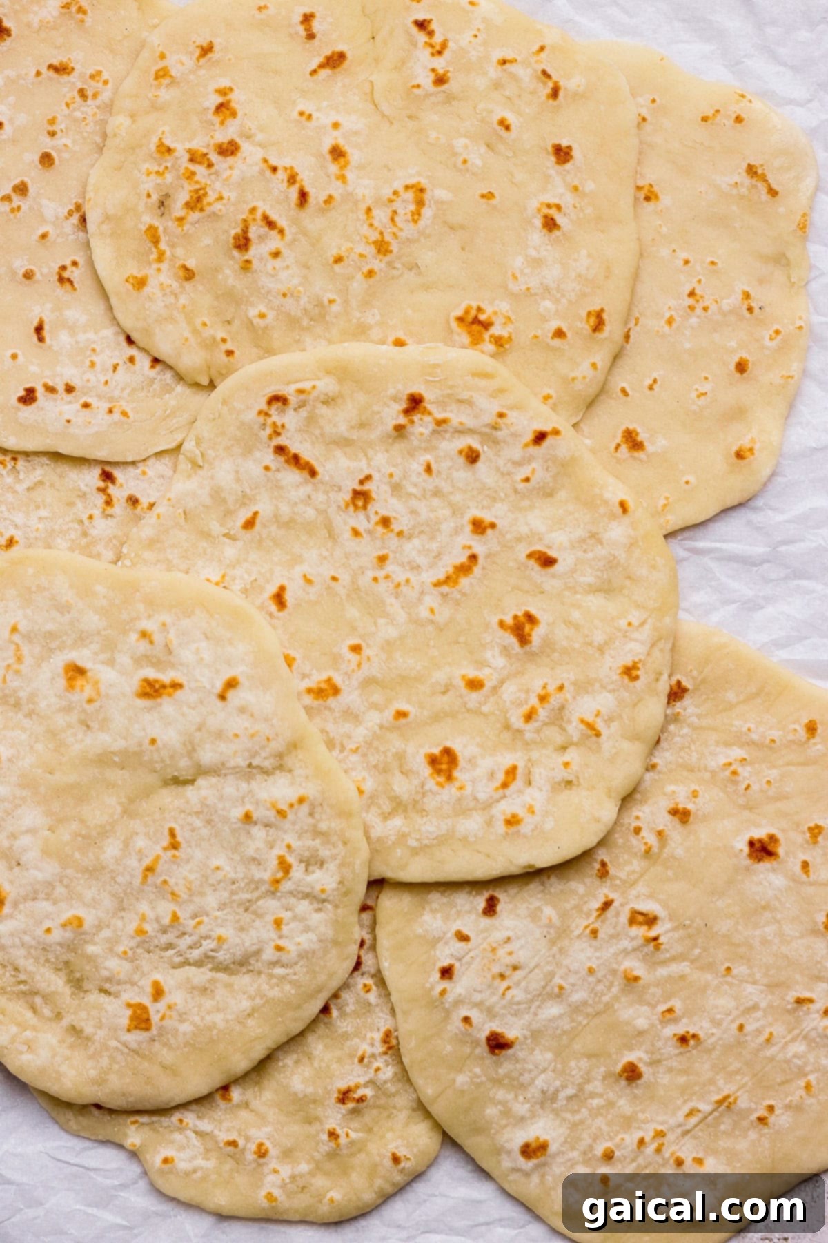 A close-up shot of several pieces of perfectly cooked flatbread, overlapping slightly, showcasing their soft texture and lightly browned spots, arranged on a rustic crumpled white surface.