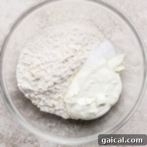 A glass bowl containing flour, white powder (possibly salt), and a dollop of plain yogurt, ready to be mixed, on a light textured surface.