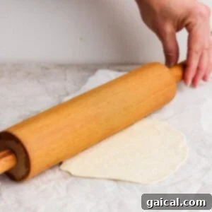 A hand holds a wooden rolling pin, meticulously rolling out a piece of dough on parchment paper, spread over a light-colored surface, aiming for thinness.