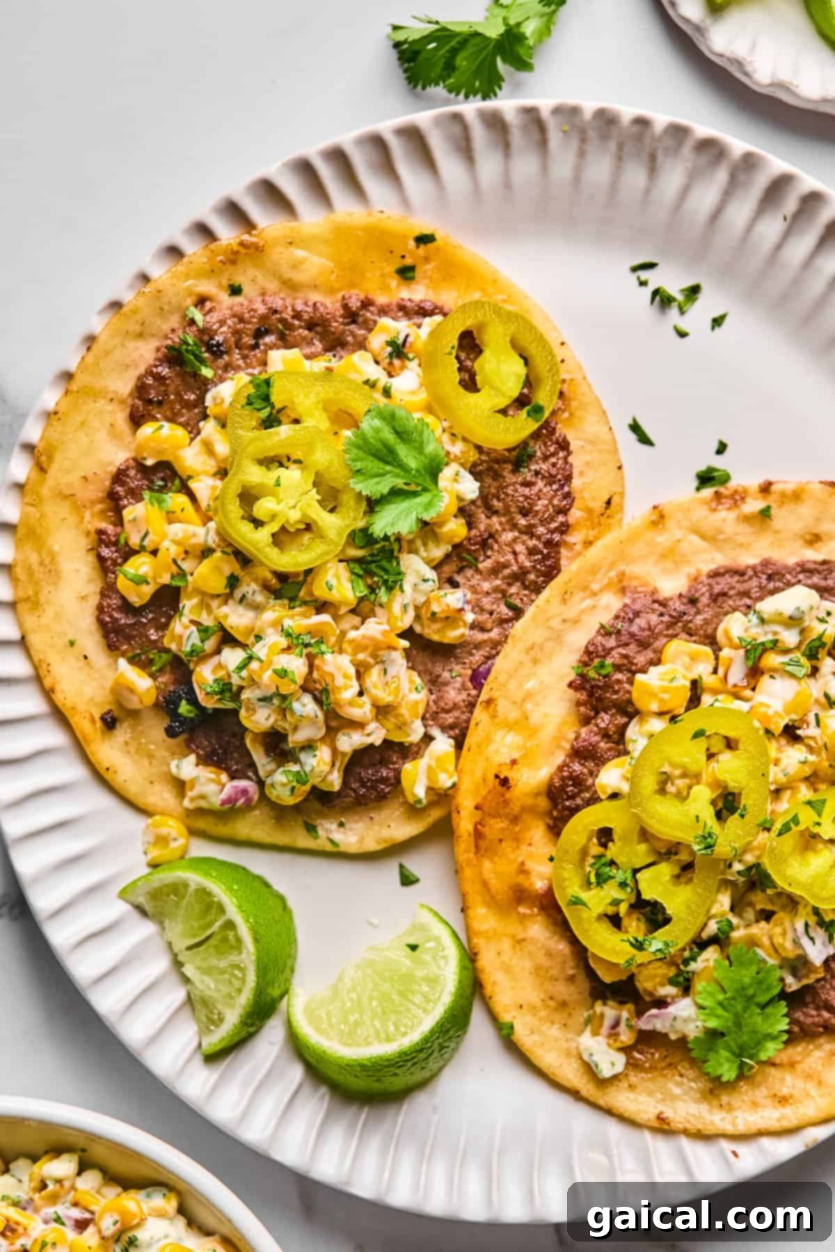 Zesty Street Corn Smash Tacos 2 Two Street Corn Smash Tacos on a white plate, generously topped with seasoned ground meat, a creamy street corn salad, vibrant pickled jalapeños, and fresh cilantro, served alongside bright lime wedges.