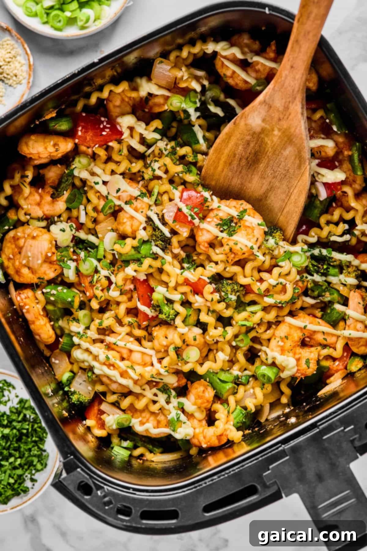 Crispy Air Fryer Shrimp and Roasted Vegetables 2 A wooden spoon stirs a mix of curly pasta, shrimp, vegetables, and herbs in an air fryer basket, with bowls of green onions and chopped herbs nearby.