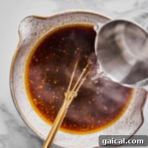 Crispy Air Fryer Shrimp and Roasted Vegetables 5 A metal whisk rests in a bowl of dark liquid while water is being poured in from a metal pitcher, all on a marble surface.