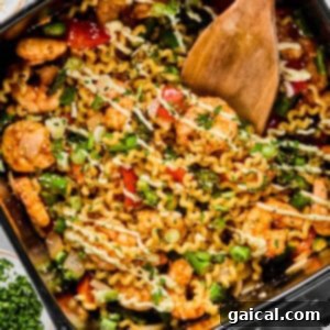 Crispy Air Fryer Shrimp and Roasted Vegetables 7 A wooden spoon stirs a mix of pasta, shrimp, vegetables, and herbs in an air fryer basket, with bowls of chopped green onions and herbs nearby.
