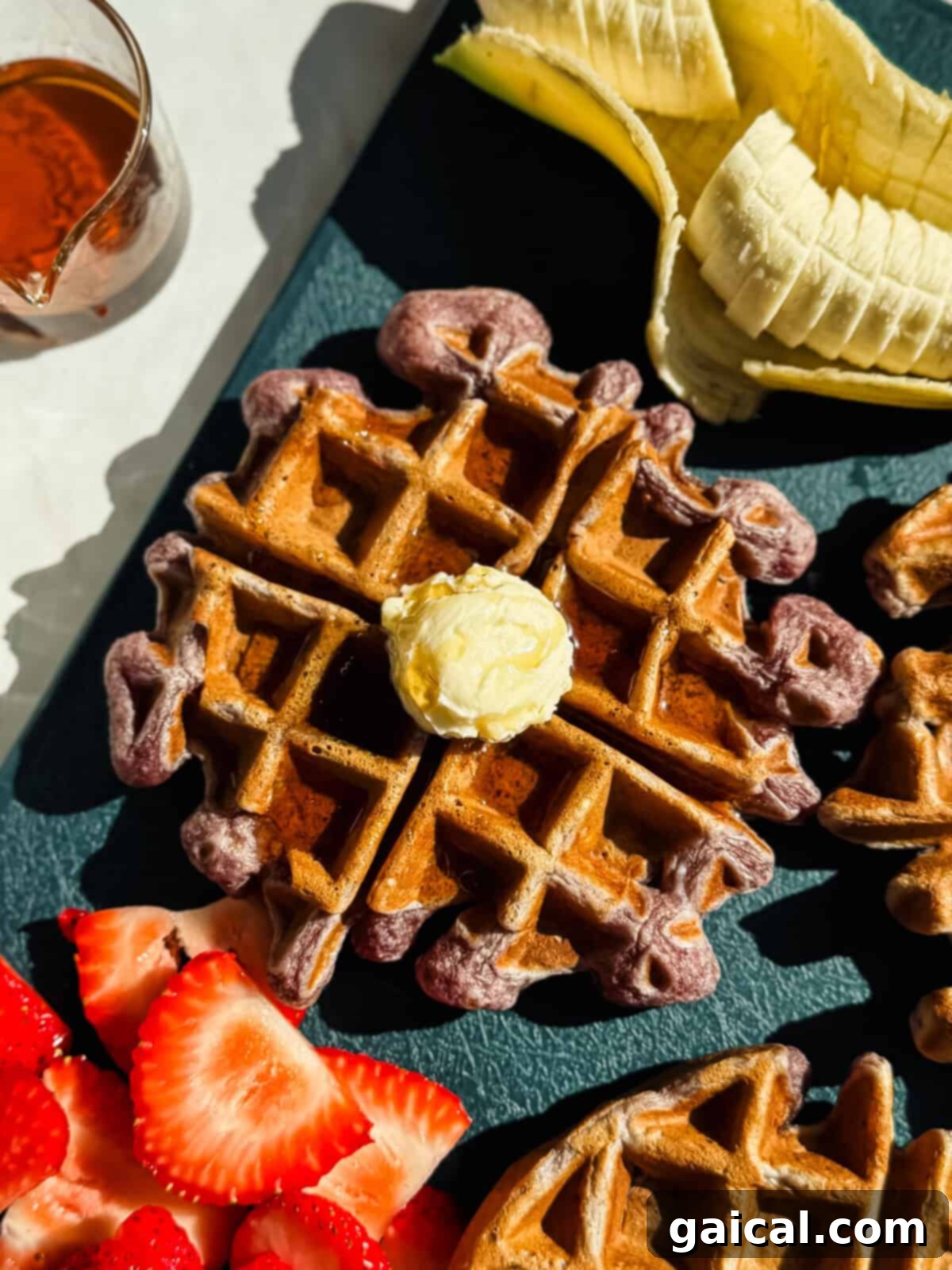 Protein-Packed Strawberry Banana Waffles 2 A waffle topped with a pat of butter is shown alongside sliced bananas, strawberries, and a small glass of syrup on a dark surface.